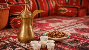 إبريق نحاسي وكؤوس بيضاء صغيرة بجانب طبق مليء بالتمر، مع خلفية سجادة مزخرفة وأثاث تقليدي.