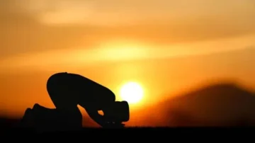 Silhouette d'une personne en prière au coucher du soleil, avec un ciel orange et des montagnes en arrière-plan.