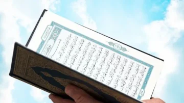 Un homme tient un livre sacré ouvert, avec des pages écrites en arabe, sur fond de ciel bleu et nuages. Ce livre représente le Coran, un texte central de la foi islamique.