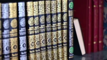 Collection de livres anciens sur une étagère, incluant plusieurs volumes reliés en cuir noir ornés de motifs dorés et de titres en arabe. À côté, un livre de couleur claire sans couverture visible.