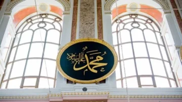 Inscription en calligraphie arabe du nom du prophète Mohammed dans un cadre ovale, suspendue entre de grandes fenêtres. Éclairage naturel illuminant l'intérieur, mettant en valeur l'art et l'architecture islamiques.