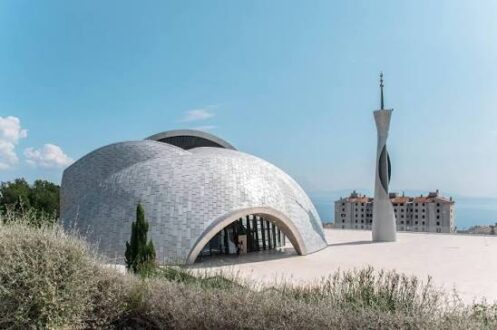 مبنى حديث بتصميم معمارى فريد، يضم قبة كبيرة ومئذنة أنيقة. يقع في منطقة مرتفعة مع مناظر طبيعية، وتحيط به الأعشاب والنباتات. السماء زرقاء وصافية، مما يضيف جمالية للمشهد.