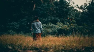 Person de dos marchant dans un champ de haute herbe, entouré d'arbres verdoyants. Lumière douce et ambiance calme, évoquant la nature et la solitude.