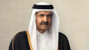 صورة لرجل يرتدي الزي التقليدي القطري، يتضمن عباءة وقبعة، مع تعبير جاد. الخلفية بسيطة وتضفي طابعاً رسمياً.