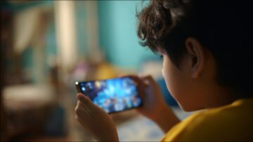طفل يجلس في غرفة مضاءة خافتة، يحمل هاتفًا ذكيًا في يده. يركز على شاشة الهاتف، حيث تعرض لعبة مثيرة. تظهر ملامح التركيز على وجهه في حين تنعكس الأضواء من الشاشة على عينيه.
