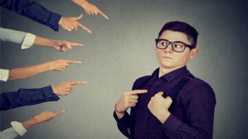 رجل يرتدي نظارات وقميص مزخرف، يبدو متوترًا وهو ينظر إلى عدة أيدي تشير إليه بأصابعها. تعبير وجهه يظهر القلق أو الإحراج، والخلفية رمادية. الصورة تعكس مشاعر الاتهام أو الإدانة.