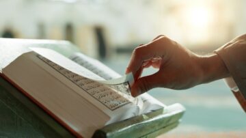 يد تُقلب صفحات القرآن الكريم في مسجد، مع خلفية مضيئة تظهر أجواء الروحانية والسكينة.