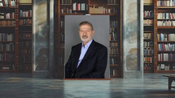 رجل في بدلة رسمية يجلس أمام خلفية مكتبة تحتوي على رفوف مليئة بالكتب. الإضاءة تبرز ملامحه، مما يضفي طابعًا رسميًا ومهنيًا على الصورة.