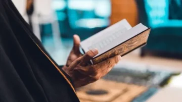 Person tenant un livre ouvert, avec un accent sur les pages, dans un environnement intérieur chaleureux et confortable. Le décor comprend des canapés et des éléments de style moderne. Idéal pour des thèmes liés à la lecture, la méditation ou la spiritualité.