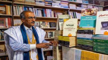 رجل مسن يرتدي جلابية وعمامة، يقف بجانب رفوف مليئة بالكتب في مكتبة. تظهر على الرفوف عناوين كتب متنوعة باللغة العربية، بما في ذلك كتب عن الثقافة والأدب. تعكس الصورة أجواء المعرفة والتعلم في بيئة ثقافية غنية.
