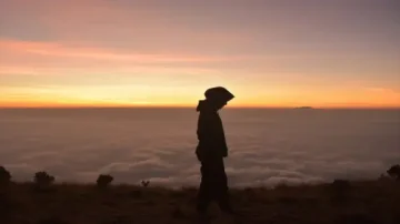 Silhouette d'une personne marchant sur un sommet avec un lever de soleil éclatant en arrière-plan, entourée de nuages.