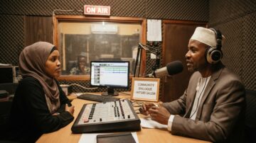 A candid shot inside a modest community radio station in East Africa. Hitsri Salem, wearing a patterned shirt and a warm smile, speaks closely into a professional broadcast microphone.