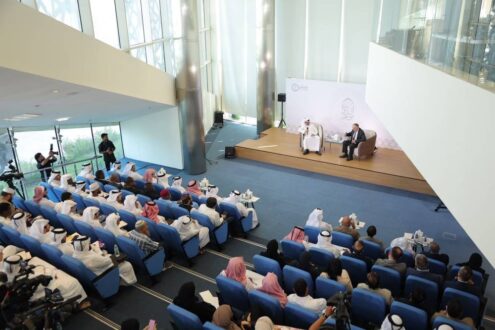 ندوة في قاعة حديثة مع جمهور كبير من الحضور يرتدون ملابس تقليدية، يتحدث فيها متحدثان على المنصة. تركز الإضاءة الطبيعية على المتحدثين بينما يجلس الحضور على كراسي زرقاء في صفوف منظمة.