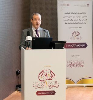 رجل يتحدث في مؤتمر حول القرآن والدراسات الإنسانية، يقف أمام منصة تحتوي على شعار المؤتمر. خلفه لافتة تحمل تفاصيل الحدث.