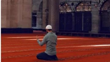 Homme en prière dans une mosquée, vêtu d'une chemise grise et d'un bonnet blanc, sur un tapis rouge. L'intérieur de la mosquée présente des colonnes majestueuses et des détails architecturaux.
