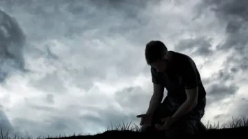 Homme en silhouette accroupi sur un sol herbeux, les mains tendues vers le sol, sous un ciel nuageux et sombre. Évoque des thèmes de désespoir et de réflexion.