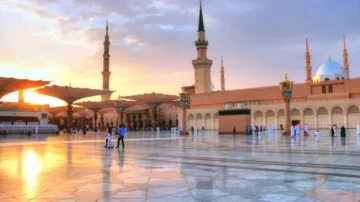 Vue panoramique de la mosquée du Prophète à Médine au coucher du soleil, avec des minarets et des parasols déployés. Des visiteurs se déplacent sur le sol en marbre, créant une ambiance paisible et spirituelle.