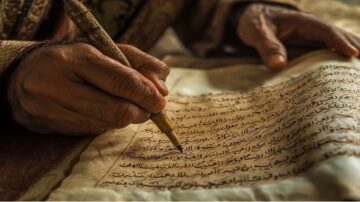 يد تمسك بقلم وتكتب على ورقة قديمة مكتوبة باللغة العربية، تظهر تفاصيل النص والخط التقليدي. الخلفية غير واضحة، مما يبرز اليد والأداة المستخدمة في الكتابة.