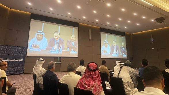 اجتماع في قاعة مؤتمرات مع شاشة عرض كبيرة تعرض متحدثين على المنصة. الحضور يرتدون ملابس تقليدية وعصرية، مع التركيز على النقاشات والأفكار المطروحة.