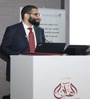 رجل يرتدي بدلة يتحدث في مؤتمر بحضور جمهور. خلفه شاشة عرض تحتوي على نص باللغة العربية، تتعلق بالأبحاث والدراسات.