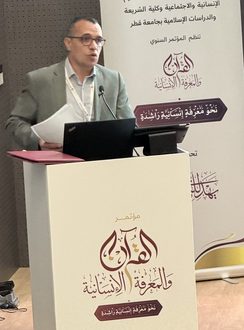 رجل يتحدث في مؤتمر حول القضايا الإنسانية والاجتماعية، مع خلفية تحمل شعار المؤتمر. يظهر المتحدث وهو يرتدي بدلة، ويقف أمام منصة تحمل اسم المؤتمر.