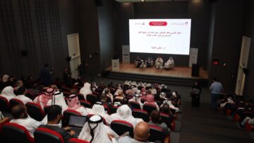 ندوة في قاعة مؤتمرات مع جمهور كبير من الحضور، يتضمن panel من المتحدثين على المسرح. الشاشة تعرض معلومات حول الموضوع، مع وجود رجال ونساء يرتدون الملابس التقليدية.