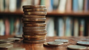 عملات معدنية مكدسة على طاولة خشبية، مع عملات متناثرة حولها، وخلفية تحتوي على رفوف كتب.