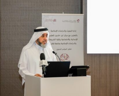 رجل يرتدي الزي التقليدي يتحدث في مؤتمر إسلامي، مع ميكروفون أمامه وشاشة تعرض شعار المؤتمر. خلفية المكان تظهر تصميمًا عصريًا.
