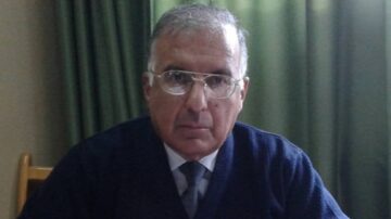 رجل مسن يرتدي نظارات وقميصًا داكنًا يجلس أمام ستائر خضراء. يبدو جادًا وهو ينظر مباشرة إلى الكاميرا، مما يسلط الضوء على ملامح وجهه.