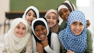 Un groupe de six jeunes filles portant des hijabs sourient joyeusement à la caméra. Elles sont assises ensemble, affichant une atmosphère de camaraderie et de bonheur. L'environnement est lumineux et accueillant, mettant en valeur la diversité culturelle.