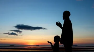 Silhouette d'un adulte et d'un enfant en train de prier sur la plage au coucher du soleil, avec des nuages dans le ciel et des reflets sur l'eau.