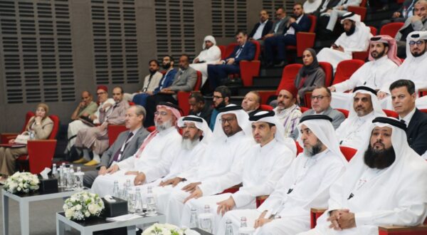 حضور مجموعة من الرجال والنساء في قاعة مؤتمرات، حيث يجلس عدد من الرجال يرتدون الزي التقليدي الإماراتي في المقدمة، بينما يظهر جمهور متنوع في الخلفية، مع التركيز على المنصة والألوان الزاهية للمقاعد.