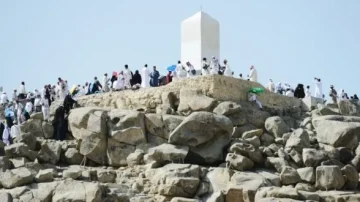 Montagne de la Miséricorde à La Mecque, avec des pèlerins en train d'explorer les rochers. Un monument blanc se dresse au sommet, entouré de visiteurs en vêtements traditionnels. Ciel clair et ensoleillé.