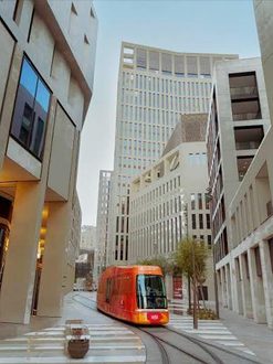 ترام جديد يجري عبر حي حديث مع مبانٍ شاهقة. يظهر التصميم المعماري العصري والطرق المعبدة، مما يعكس الحياة الحضرية المتطورة.