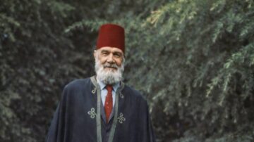 رجل مسن يرتدي الزي التقليدي مع عمامة حمراء ولحية بيضاء، يقف في بيئة طبيعية مع أشجار خضراء في الخلفية.