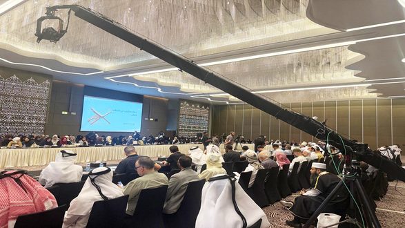 اجتماع حاشد في قاعة مؤتمرات مع مجموعة متنوعة من الحضور، بما في ذلك رجال ونساء يرتدون ملابس تقليدية. يوجد شاشة عرض خلف المنصة تعرض محتوى باللغة العربية. في المقدمة، يوجد ذراع كاميرا طويل يلتقط الحدث. الأجواء تعكس تفاعلًا حيويًا.