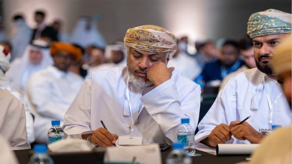 رجل يرتدي العمانية التقليدية يجلس في مؤتمر، يكتب ملاحظات بتركيز. في الخلفية، مجموعة من الحضور في ملابس متنوعة. زجاجات مياه على الطاولة.