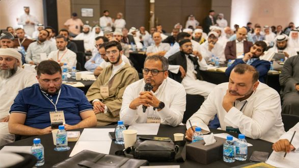 حضور كبير في مؤتمر، رجال ونساء يجلسون في قاعة، بعضهم يتحدث وآخرون يستمعون. الطاولات تحتوي على زجاجات مياه وأدوات مكتبية، مع بعض المشاركين يركزون على المتحدث.