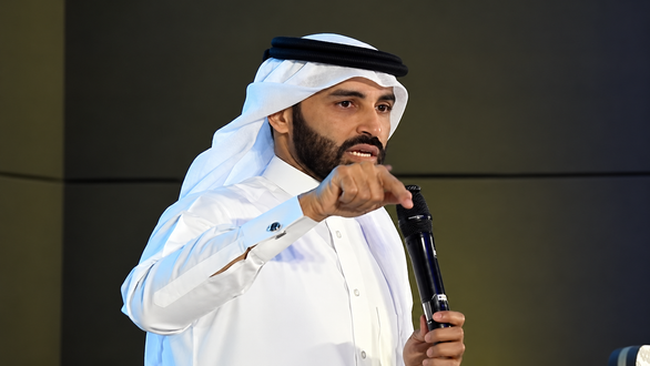 رجل عربي يرتدي الزي التقليدي، يتحدث بحماسة أمام جمهور، ممسكًا بميكروفون. خلفية الحدث تظهر ألوان دافئة، مما يبرز أهمية الرسالة المقدمة.