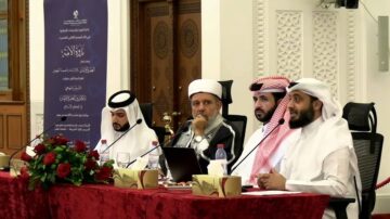 رجلان يرتديان الملابس التقليدية العربية يجلسان على طاولة مزينة بالورود، مع وجود رجل ثالث يحمل ميكروفون، في حدث رسمي. خلفهم لافتة تحمل نصوصًا باللغة العربية.