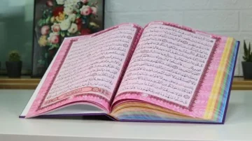 Livre ouvert sur une table, montrant des pages de texte en arabe. Le livre présente un design coloré avec des marges décoratives. En arrière-plan, des plantes en pot et une composition florale ajoutent une touche esthétique.