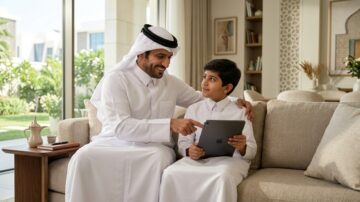رجل عربي يرتدي الزي التقليدي يجلس مع طفل في غرفة معيشة حديثة. كلاهما يتطلع إلى جهاز آيباد، ويبدو عليهما الفرح والتركيز. خلفية تحتوي على نوافذ واسعة تطل على حديقة.