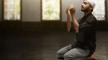 Homme en prière dans un espace intérieur sombre, genoux au sol, mains levées vers le ciel, lumière douce filtrant à travers des fenêtres. Atmosphère de sérénité et de dévotion.